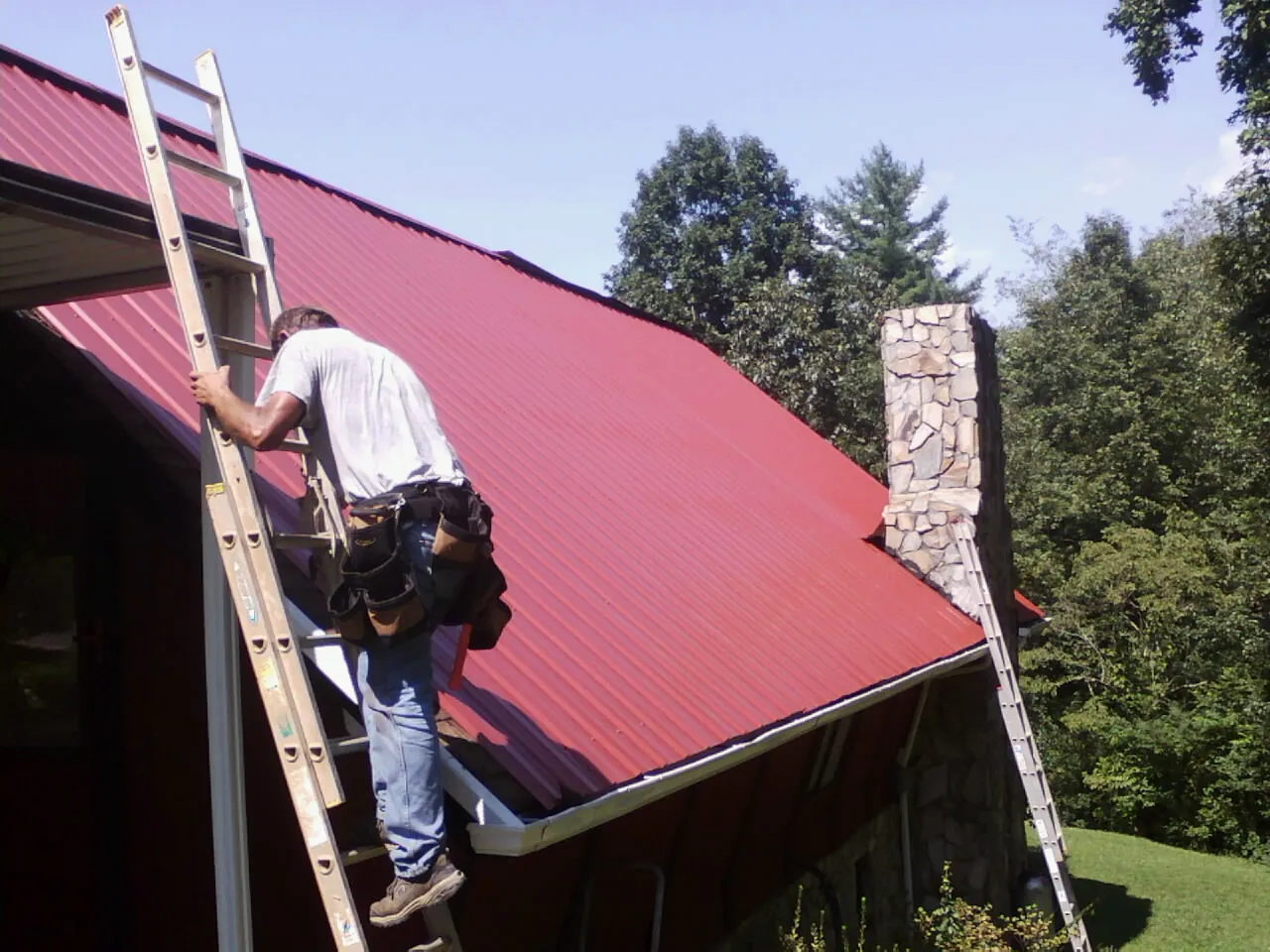 Roofer on a ladder beside a red metal roof and stone chimney — Soffit & Fascia Repair by Roofing Brothers in Rushville